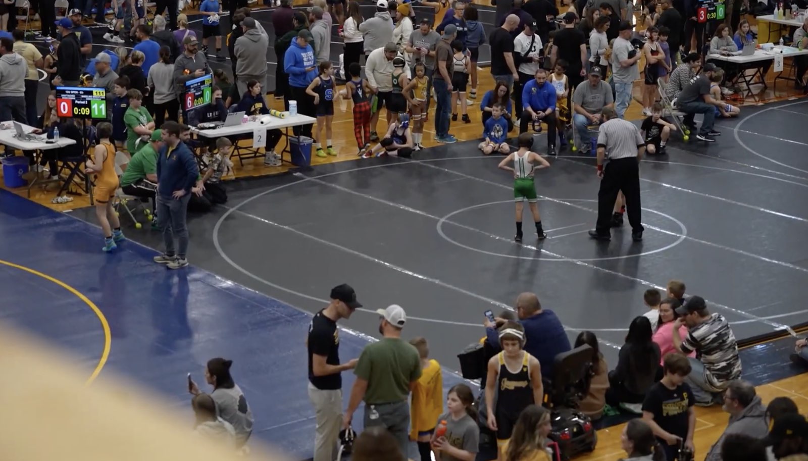 A youth tournament floor from above: three mats running, scoreboards showing live scores, tables of officials, and families around the edge.